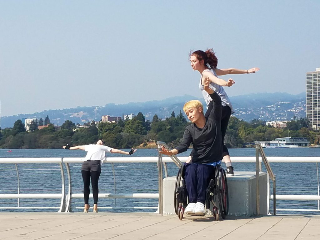 Three disabled and non-disabled dancers improvise at Lake Merritt in Oakland, CA. Jongchul is a wheelchair user with blond hair, he lifts to arms up like a bird in flight, while Kelly stands on a platform behind him and mirrors is gesture. A third dancer leans on a railing behind them with their arms outstretched horizontally.