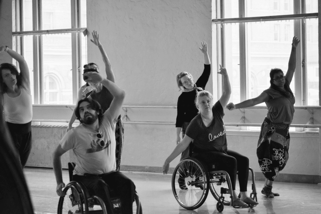 Disabled and non-disabled dancers take class; performing a floor combination in unison with one upper arm raised in a curve. 