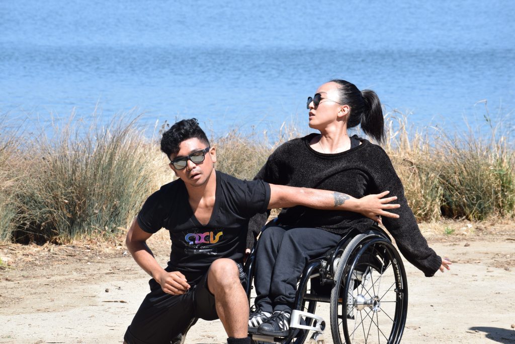 AJ crouches besides Julie, who is a wheelchair user, as they dance with Lake Merritt in the background