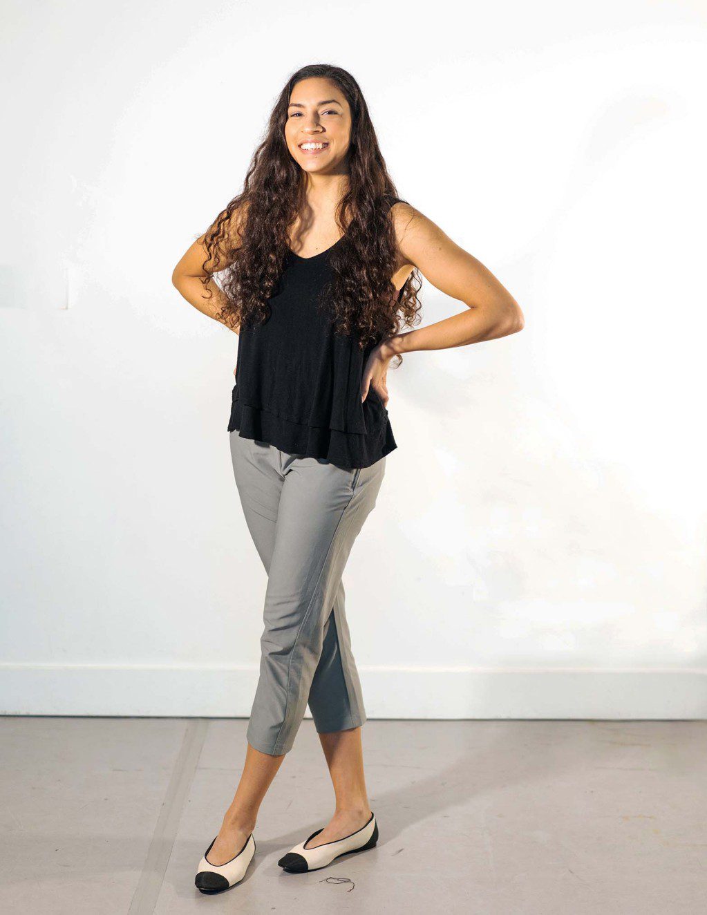 Zara smiles for the camera; she has long brown curly hair, brown eyes and skin, wearing a black top, olive green pants and ballet flats. She poses with two hands on her hips and legs crossed.