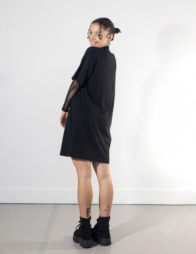 Alaja poses for the camera; they have black/caucasian mix melanated skin, short brown curly hair that's braided, and big brown eyes. They are wearing a black dress with sheer sleeves, black shoes and silver jewelry.