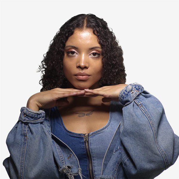 Alaja poses for the camera; they have black/caucasian mix melanated skin, short brown curly hair, and big brown eyes. They are wearing a denim jacket and tank top.