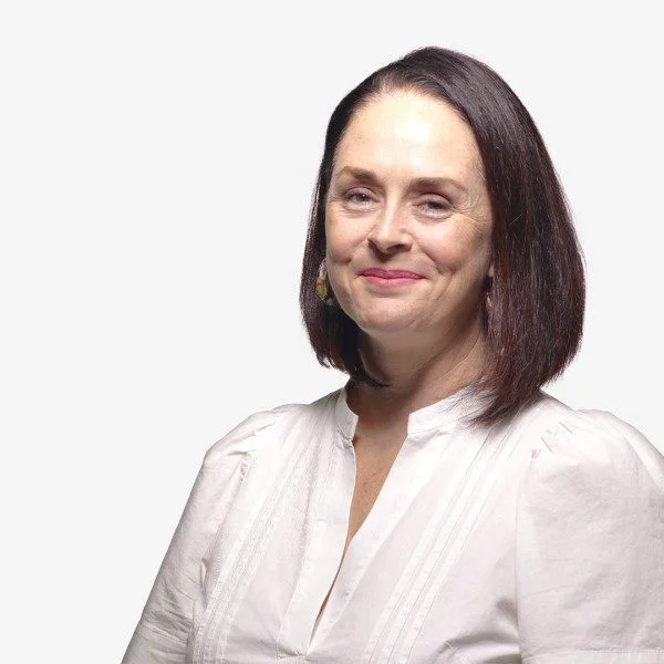 Danae Rees smiles for the camera. She is a white presenting woman with pale skin, medium length dark brown hair, and brown eyes. She is tall and slightly curve. She wears a crisp white top with dangly earrings.