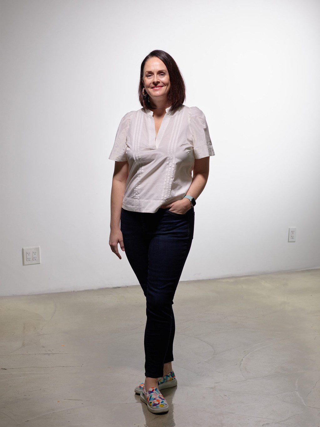 Danae Rees smiles for the camera. She is a white presenting woman with pale skin, medium length dark brown hair, and brown eyes. She is tall and slightly curve. She wears a crisp white top with dangly earrings and blue jeans.