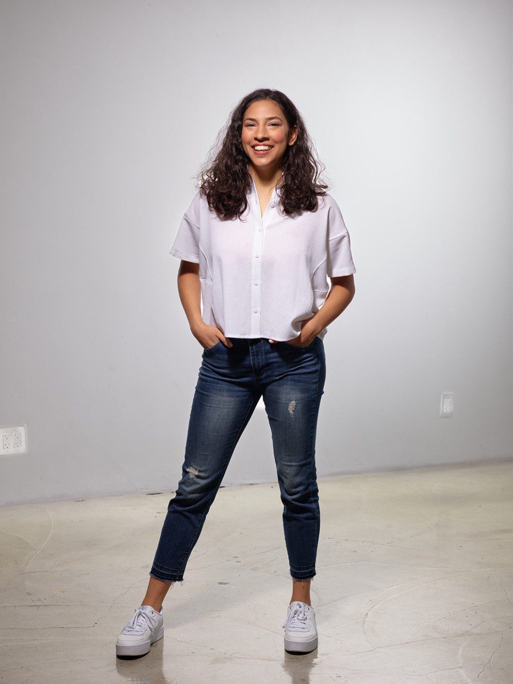 Zara poses for the camera; she has long brown curly hair, brown eyes and skin, wearing a white top and blue jeans, with her hands in her pockets.