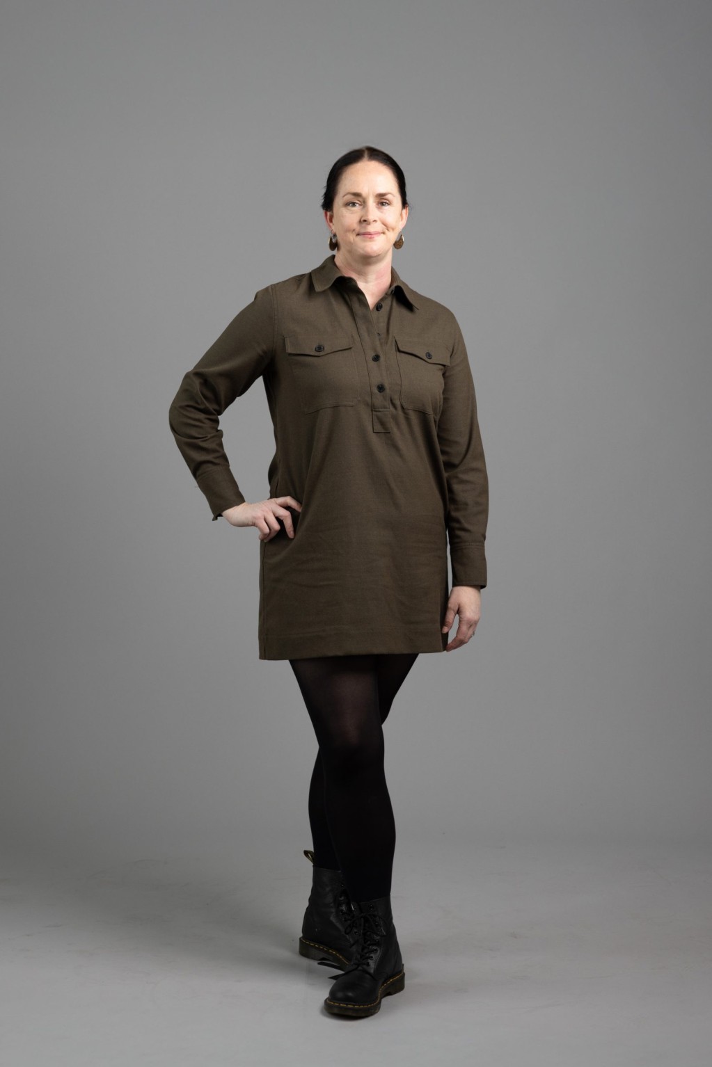 Danae smiles for the camera, with a hand on her hip. She is a white woman with black hair tied in braids; wearing an olive green dress and gold earrings.