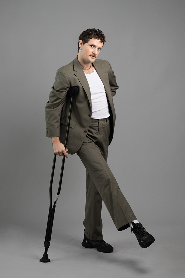 2025AXIS_RobSuguitan_Octavia_Portrait Octavia poses for a full-body portrait. They are a white person with short curly brown hair and a mustache, wearing silver jewelry and a vintage grey suit. They support their body weight with one foot and one black crutch; their other leg kicks upwards.