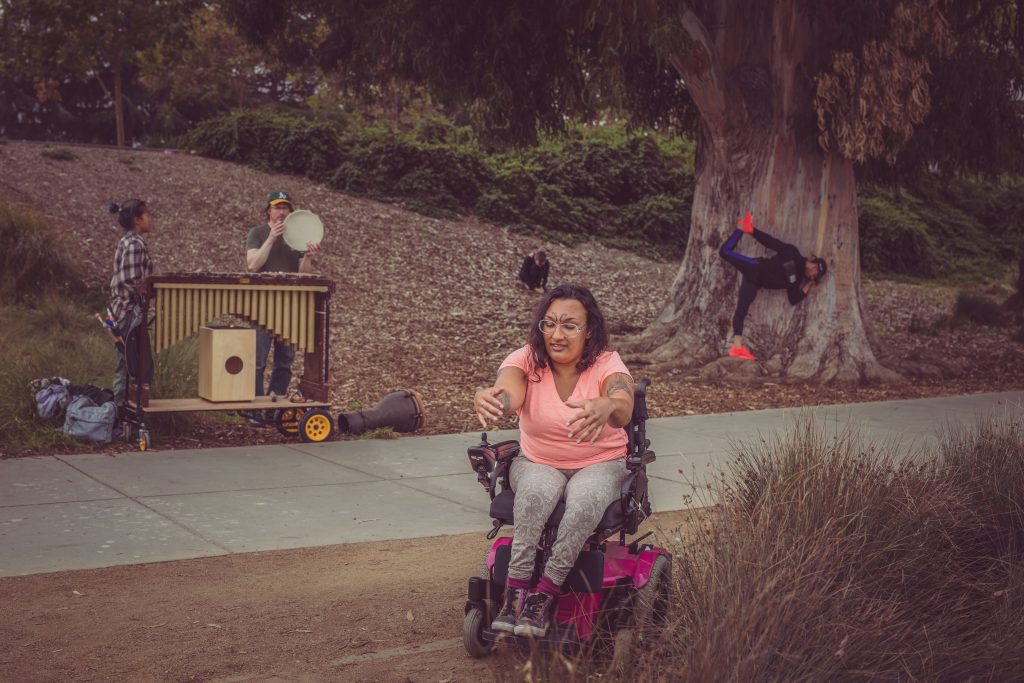 Neve, a power wheelchair user of Sudanese descent, lifts two hands up while on the shores of Lake Merrit, while two dancers pose behind her alongside a musician with a large vibraphone.