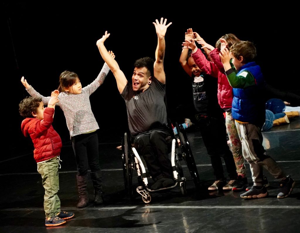 JanpiStar a latinx person who is a wheelchair user, lifts their hands up with a smile and children around them copy the gesture. 