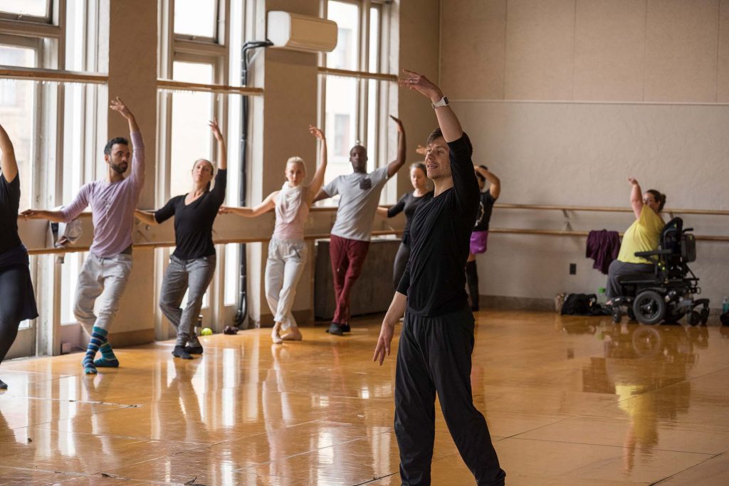 Disabled and non-disabled dancers take ballet class at the Barre, lifting upper limbs up in plié while instructor Robert Dekkers leads class. 