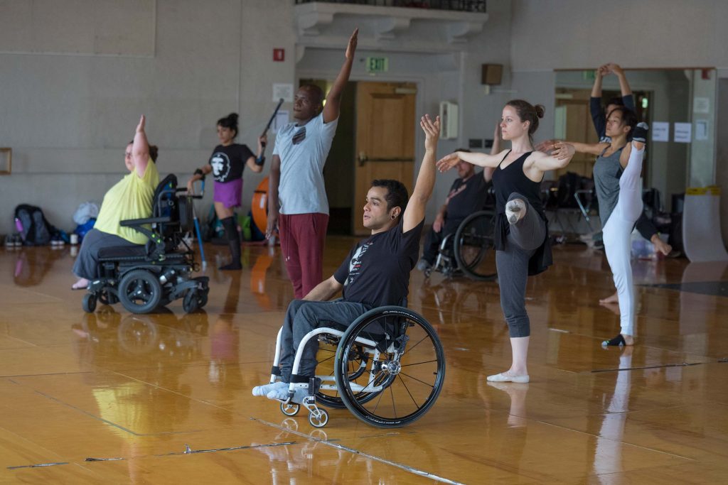 Disabled and non-disabled dancers take a ballet class together in a large dance studio with wooden floors. The dancers are practicing a grand jeté combination, using either upper or lower limbs to perform the exercise.