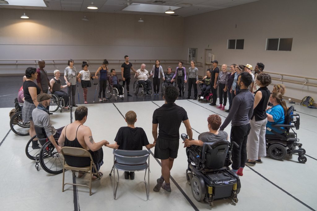 A large group of disabled and non-disabled dancers sit and stand as they hold hands in a circle in a dance studio.