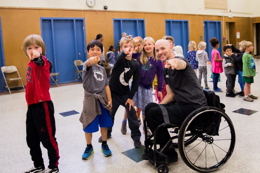 Former AXIS Dancer Dwayne Scheuneman is a white man who has a shaved head and is a wheelchair user. He smiles and poses with his finger pointing out alongside a group of young students at an assembly.