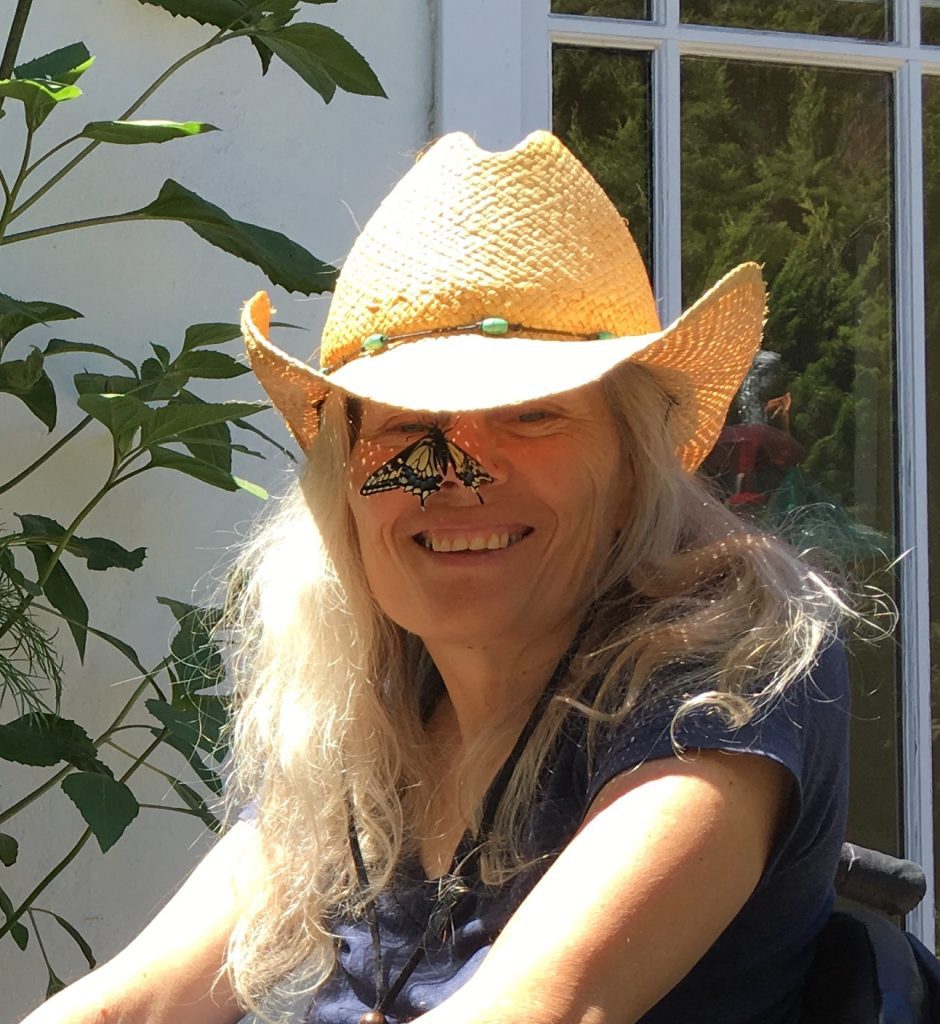 Judith Smith smiles for the camera; she is a white woman with grey blond hair who is a wheelchair user. She wears a straw cowboy hat; a yellow butterfly stops for a rest on the tip of her nose.