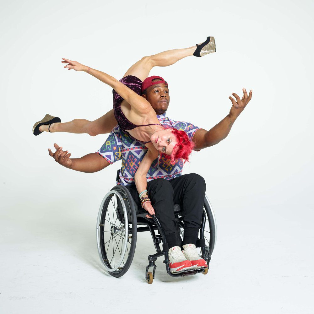 Photo by David DeSilva, dancers Sonsherée Giles and DeMarco Sleeper. Sonsherée balances the side of her torso on DeMarco’s shoulder; she extends her upper and lower limbs to the front and back while DeMarco raises his arms up. Sonsherée has bright red hair and wears a purple velvet dress; DeMarco wears a patterned T-shirt, red cap and orange & white sneakers and sits in his manual wheelchair.