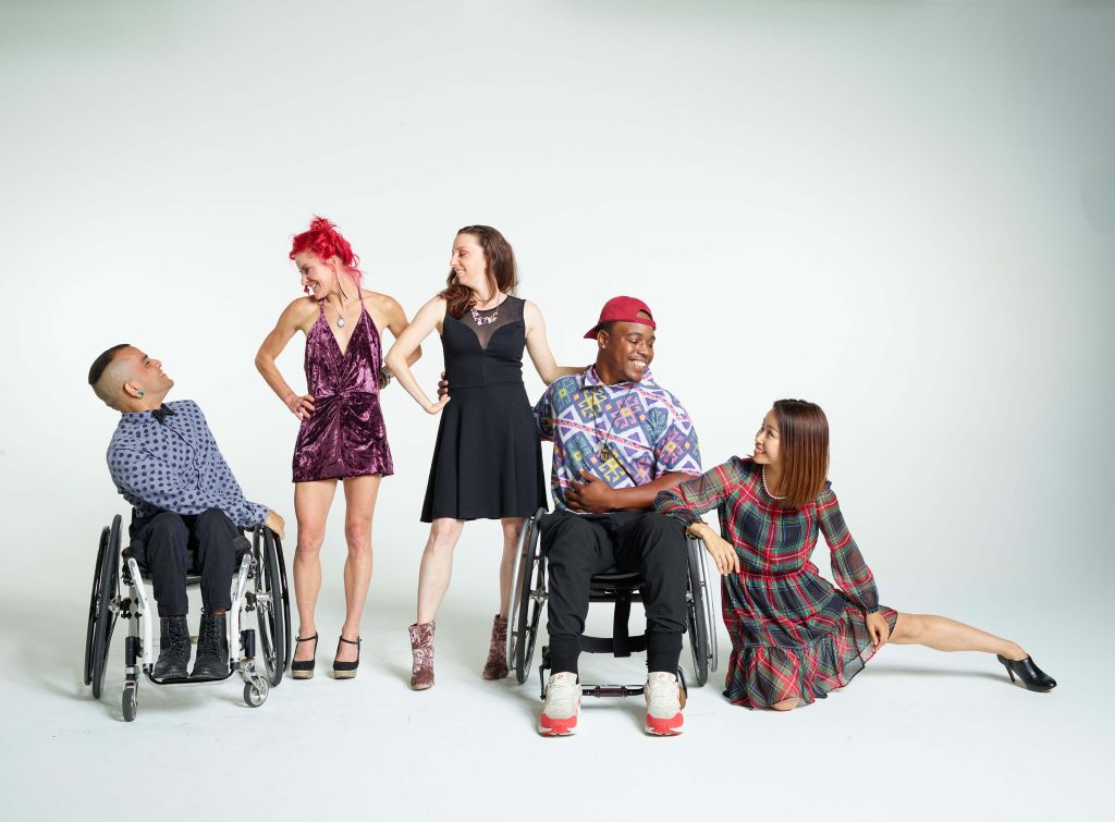 Five diverse disabled and non-disabled dancers wear brightly colored street clothes and smile as they pose together against a white background.
