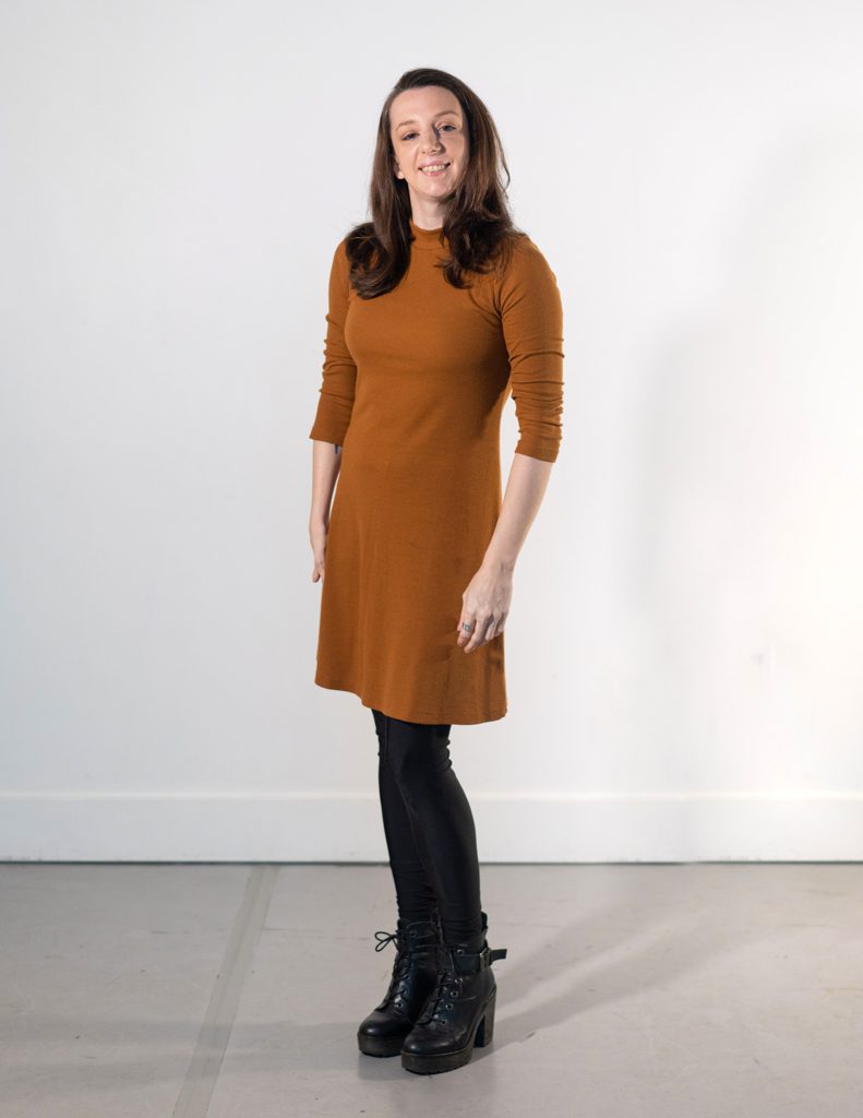 Louisa poses for the camera; she is a petite white woman in her 30s, she has an upper limb difference on the right side, auburn hair and light brown eyes. She is wearing a burnt orange turtleneck dress, black leggings and black chunky-heeled boots.