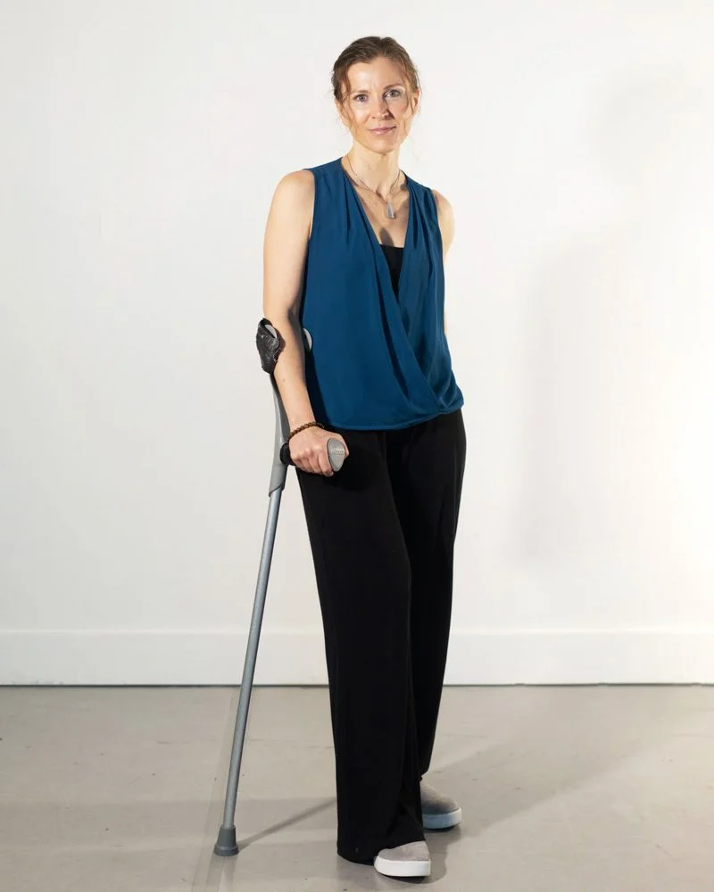 Nadia smiles for the camera. She is a cane user and a light-skinned hispanic woman with brown hair pulled back and green/blue eyes. She is wearing a black tank top with a blue, flowing sleeveless blouse on top and black, flowing pants.