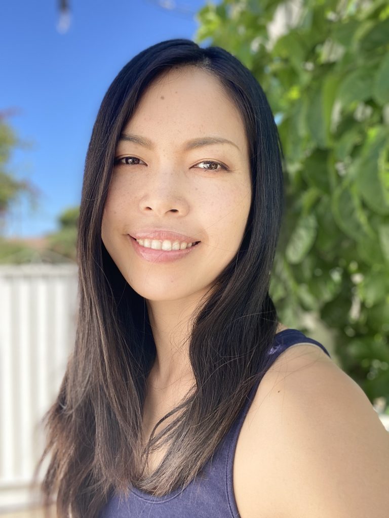 Yu is female, Asian with long black hair and brown eyes. She is wearing a navy color tank top and smiling. The background is blue sky and a green tree.