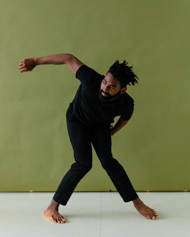Jerron Herman, a Black man with black hair and beard, tilts his body forward at an angle as his legs bend inwards. One hand reaches up and the other curves back; he is wearing all black which contrasts to an olive green wall. ,