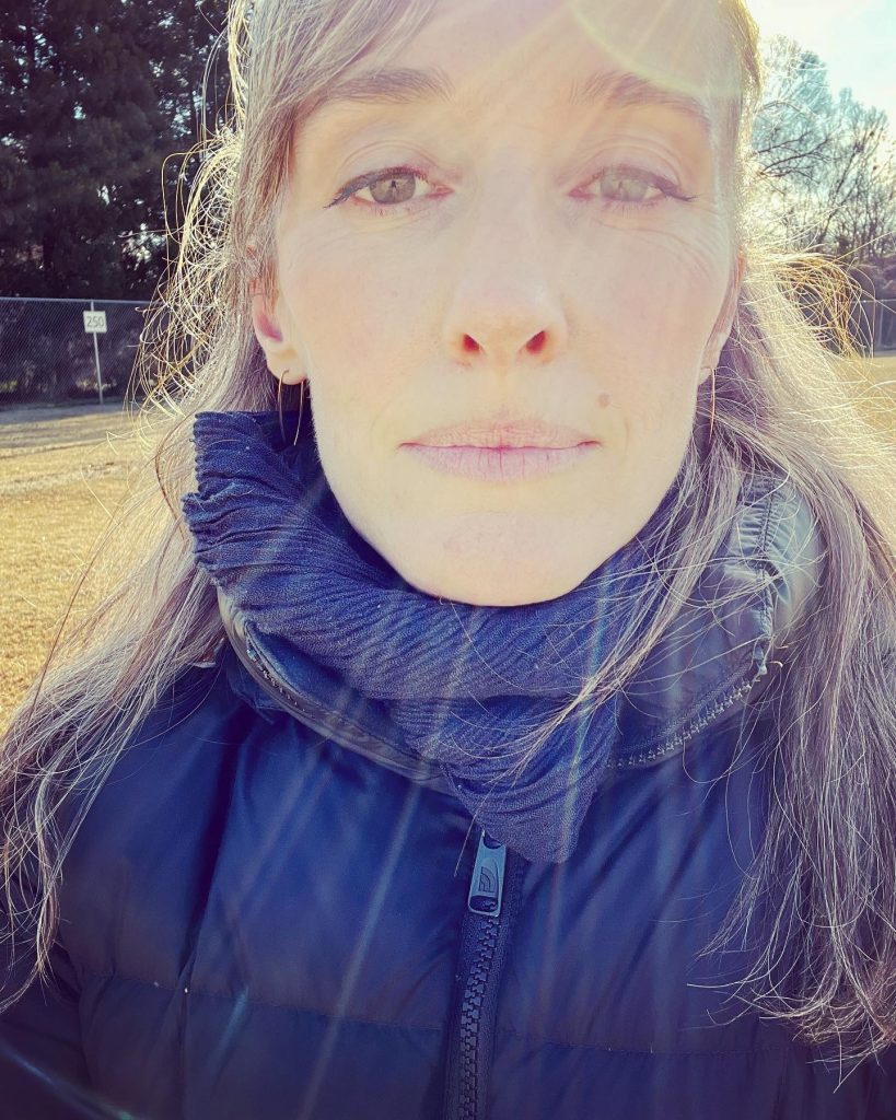 Katie looks into directly into the camera in what is clearly a selfie. Katie is a white, cis woman with long grey/blonde hair and is bundled in a navy scarf and black down jacket. She is wearing gold hoop earrings.