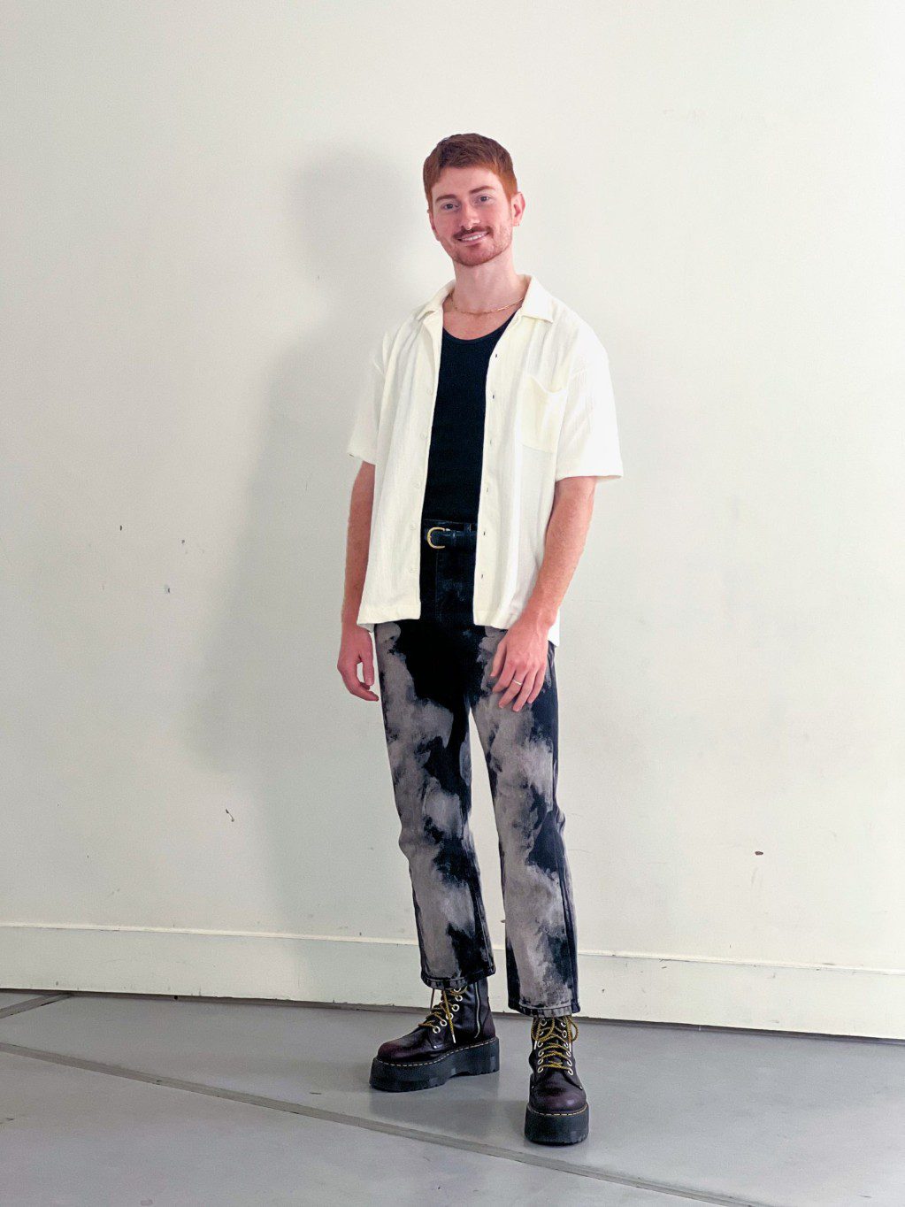 A full-body photograph of David smiling for the camera; a tall, non-disabled person with fair skin, red hair and gold jewelery with a white collared shirt, black and grey pants, and red shoes.