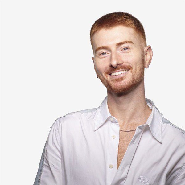 A headshot of David smiling for the camera; they are a tall, non-disabled person with fair skin, red hair and gold jewelry with a white collared shirt.