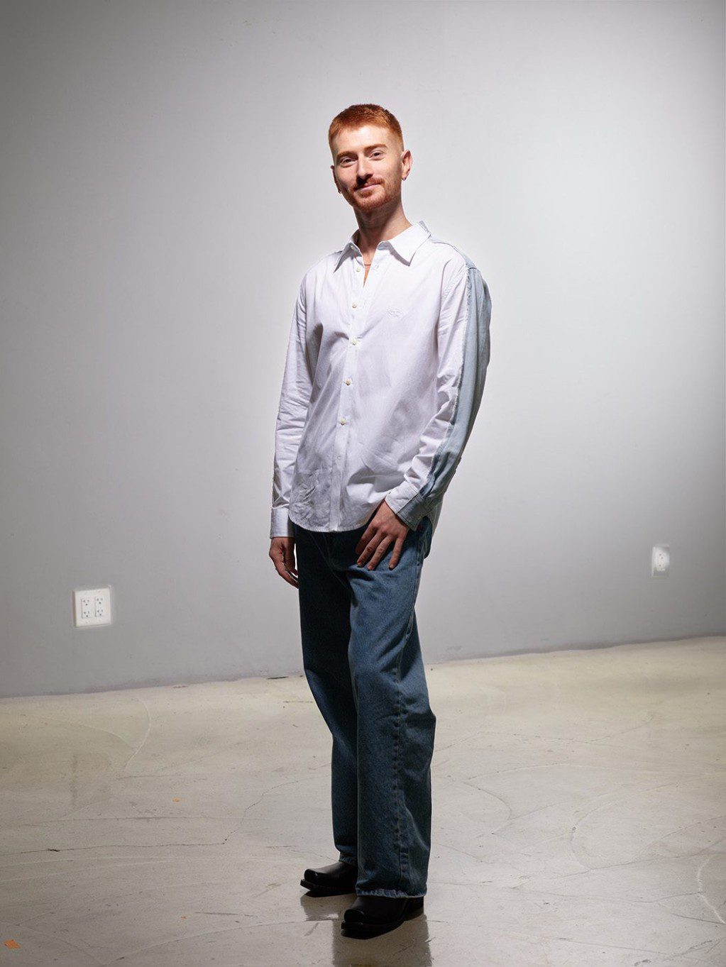 David poses for the camera; they are a tall, non-disabled person with fair skin, red hair and gold jewelry with a white collared shirt and blue jeans.
