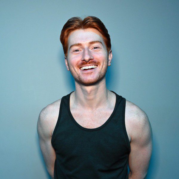 David has red hair and white skin. They are wearing a green tank top and smiling big.