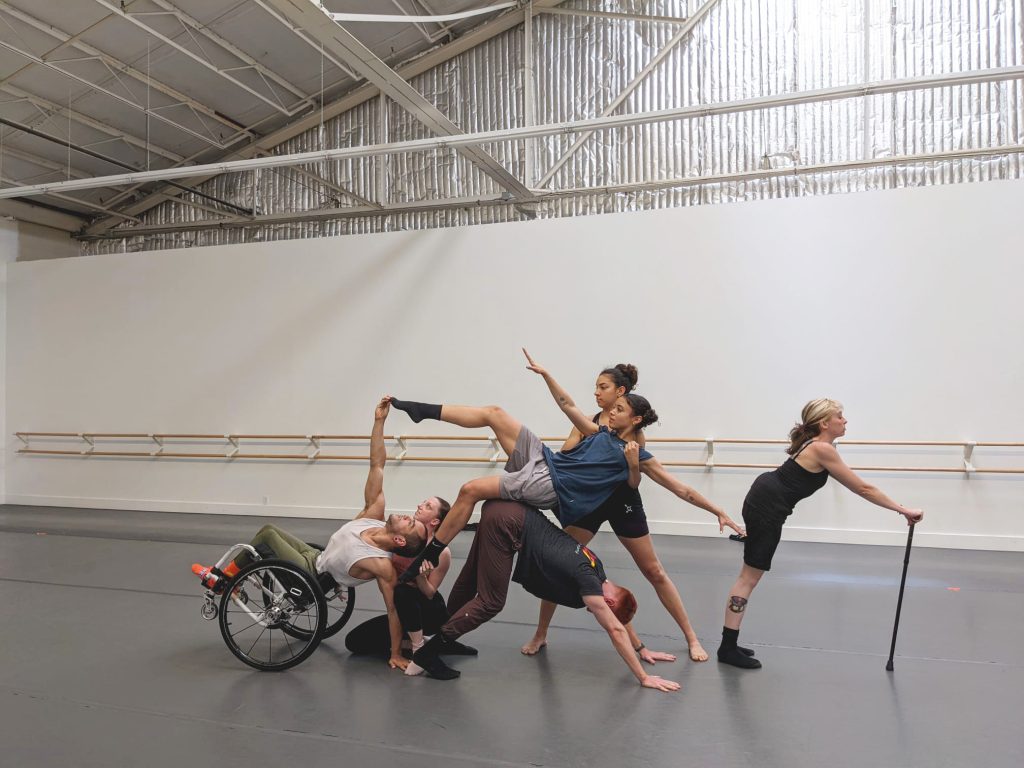Six disabled and non-disabled dancers create a tableau together in the studio. JanpiStar tilts back in their wheelchair and lifts one hand up to connect with Alaja's raised foot, who is supported by Zara while Louisa kneels beside Janpi. David forms a downward dog and Dawn leans forward on their cane to form the end of the tableau.