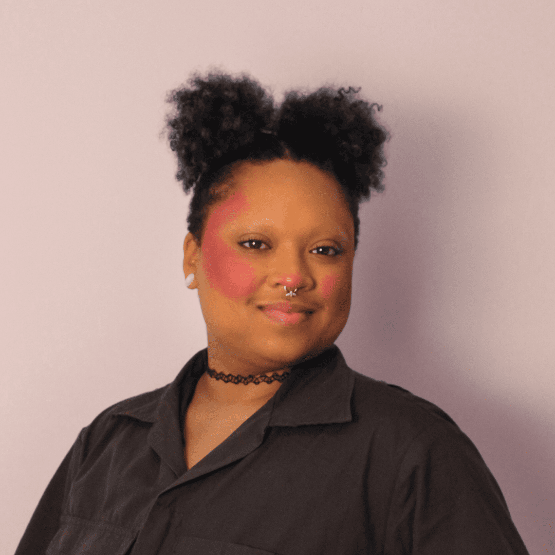 x, a Black, hypermobile, agender person stands with their face and body turned at a slight profile angle standing against a light colored wall. x has dramatic rouge on their cheeks and nose and is wearing dark gray coveralls with their dark hair in two afro puffs.