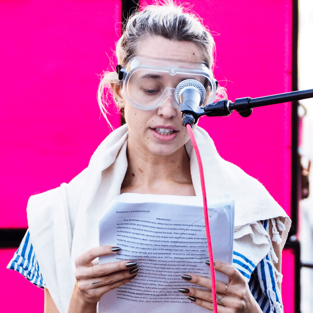 Audrey is a white presenting person with blonde hair, she wears googles and reads from a piece of paper with a pink background.