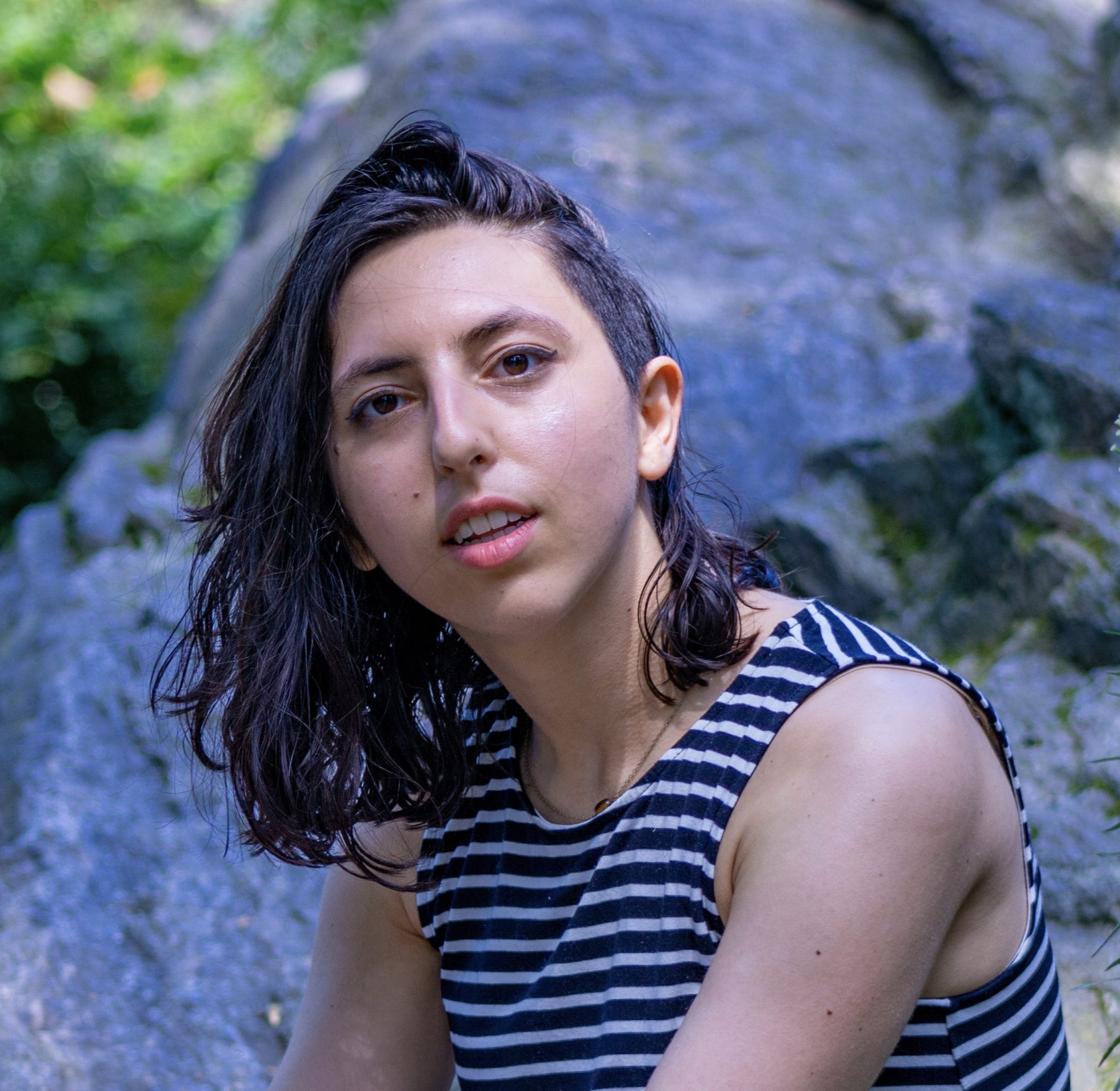 An image of Sonya from the chest up seated outdoors. Sonya is a petite white 20-something woman with layered shoulder length dark hair and dark facial features. Her body is facing slightly to the left, her head turned to the camera. There is a slight curve in her posture. Her face is alive, lips parted slightly. Her hair is flipped to the left, exposing a side shave on the right side of her head. She wears a sleeveless black and white striped top revealing muscular arms.