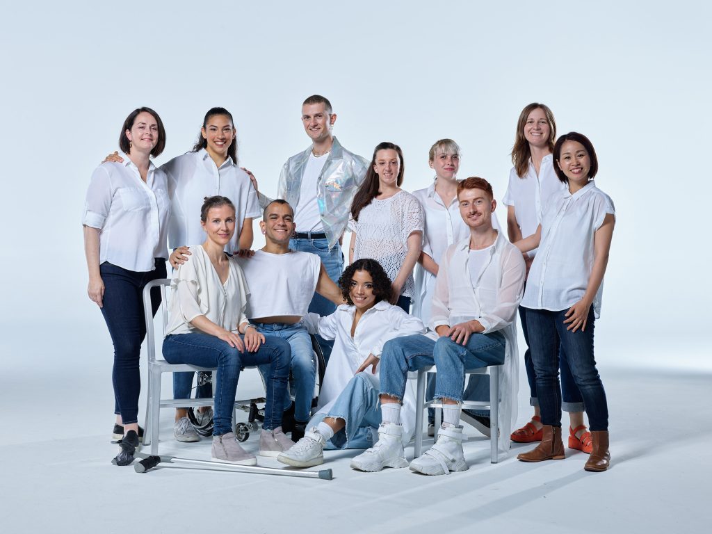 A group of 11 diverse disabled and non-disabled AXIS employees stand and sit together for a group picture. Everyone wears a white top and blue jeans, and pose against a white background.