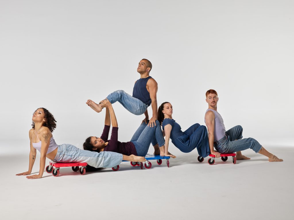 Five disabled and non-disabled dancers pose in a line with red and blue colored scooter boards. Most dancers lie and Crouch towards the floor, while JanpiStar lifts themself up from Zara's knees and hands.