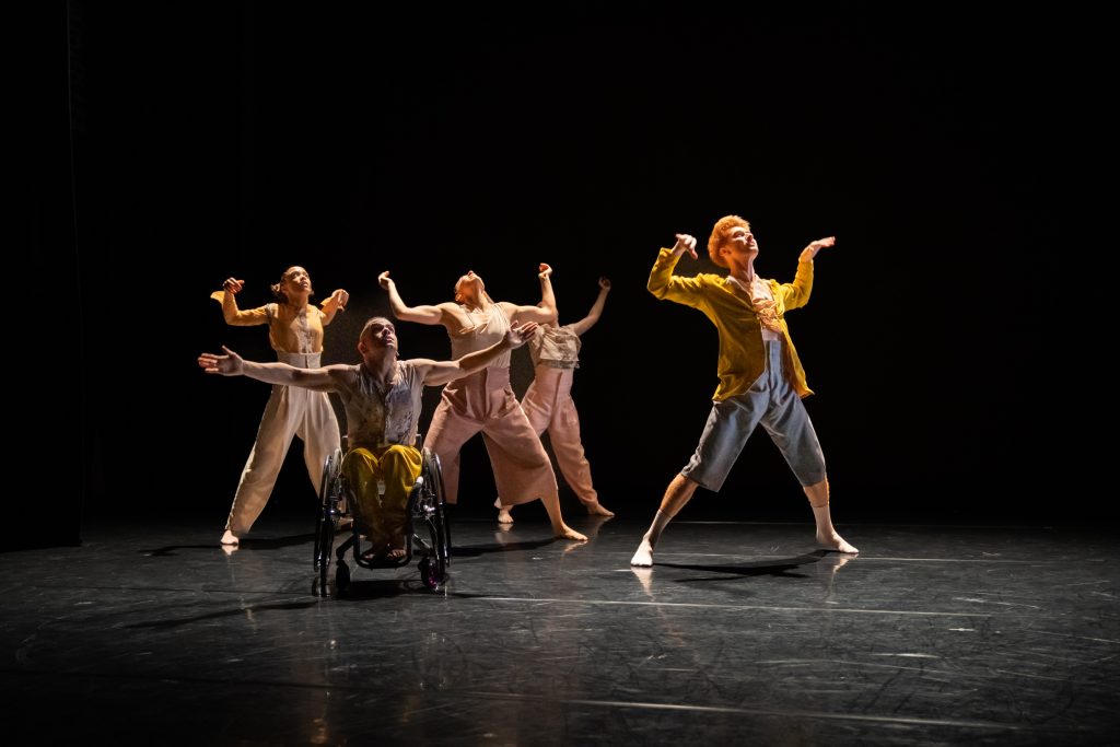 Five disabled and non-disabled dancers release their upper limbs to the ceiling after hitting their hands on their chests, which push outwards in space. The dancers wear yellow and beige costumes.