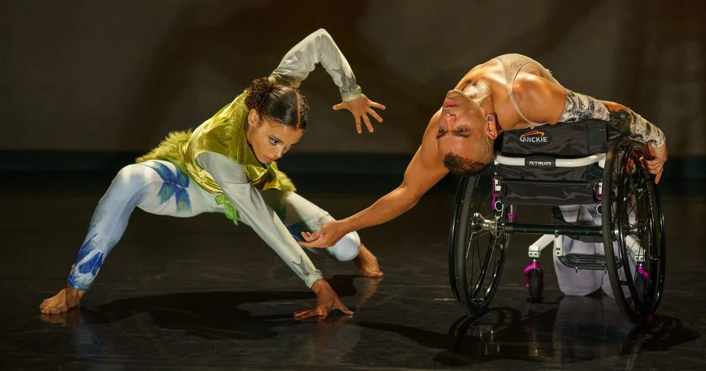 JanpiStar leans back in their wheelchair with an arched back, bringing one hand close to the floor, while Alaja Badalich crouches down in a low lunge and raises one hand up above Janpi's. Alaja wears a mossy green and turquoise costume, Janpis wears a light purple skirt and tank top.