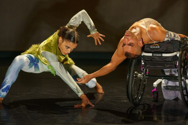 JanpiStar leans back in their wheelchair with an arched back, bringing one hand close to the floor, while Alaja Badalich crouches down in a low lunge and raises one hand up above Janpi's. Alaja wears a mossy green and turquoise costume, Janpis wears a light purple skirt and tank top.