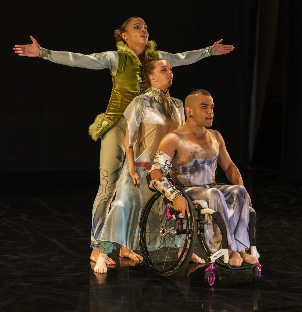 Alaja, Louisa and Janpi sit and stand in a vertical line. In the front, Janpi sits in their wheelchair, wearing a dark purple/lavender dress. Louisa plies behind them, in a turquoise gown. Alaja stands behind Louisa with arms outstretched, in a furry green vest and blue patterned unitard.