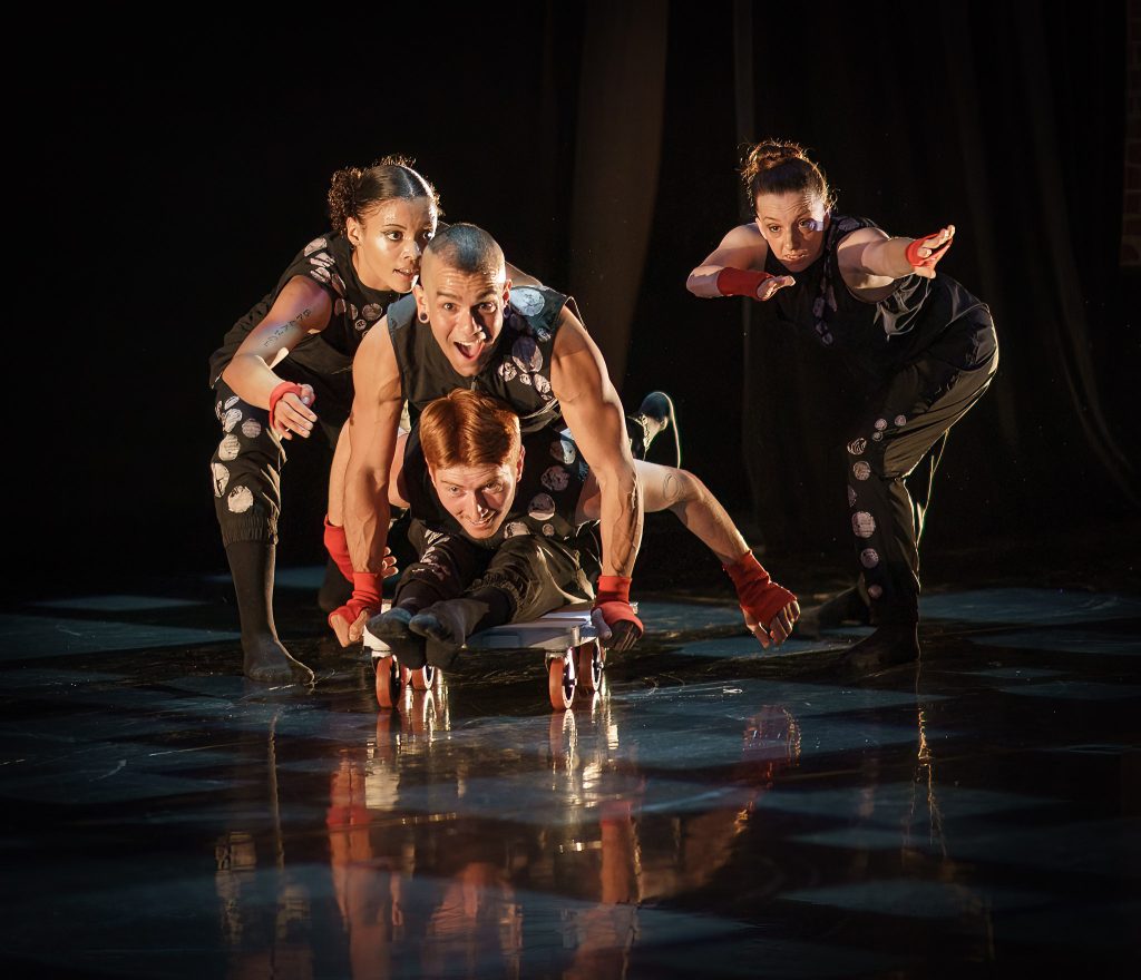 David Calhoun sits on a four-wheeled scooter, while JanpiStar lies on their back; Alaja Badalich and Louisa Mann push the dancers as they fly through space. All four dancers are smiling and wear black jumpsuits with diamond patterns.