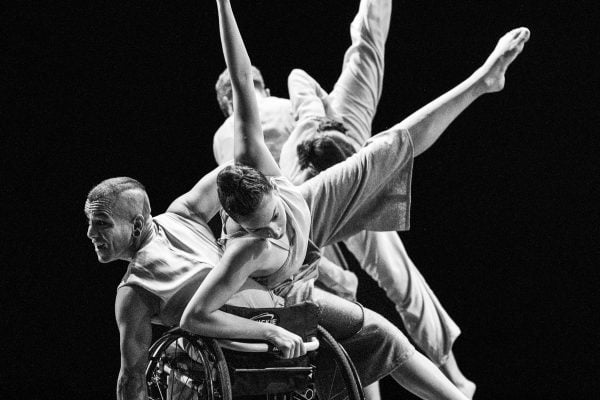 In a black and white photo, Janpi tilts their torso to the left in their wheelchair, while Zara balances in a side tilt on Janpi's body & chair with two legs and one arm extended outwards. David and Alaja perform the same movements behind them both.