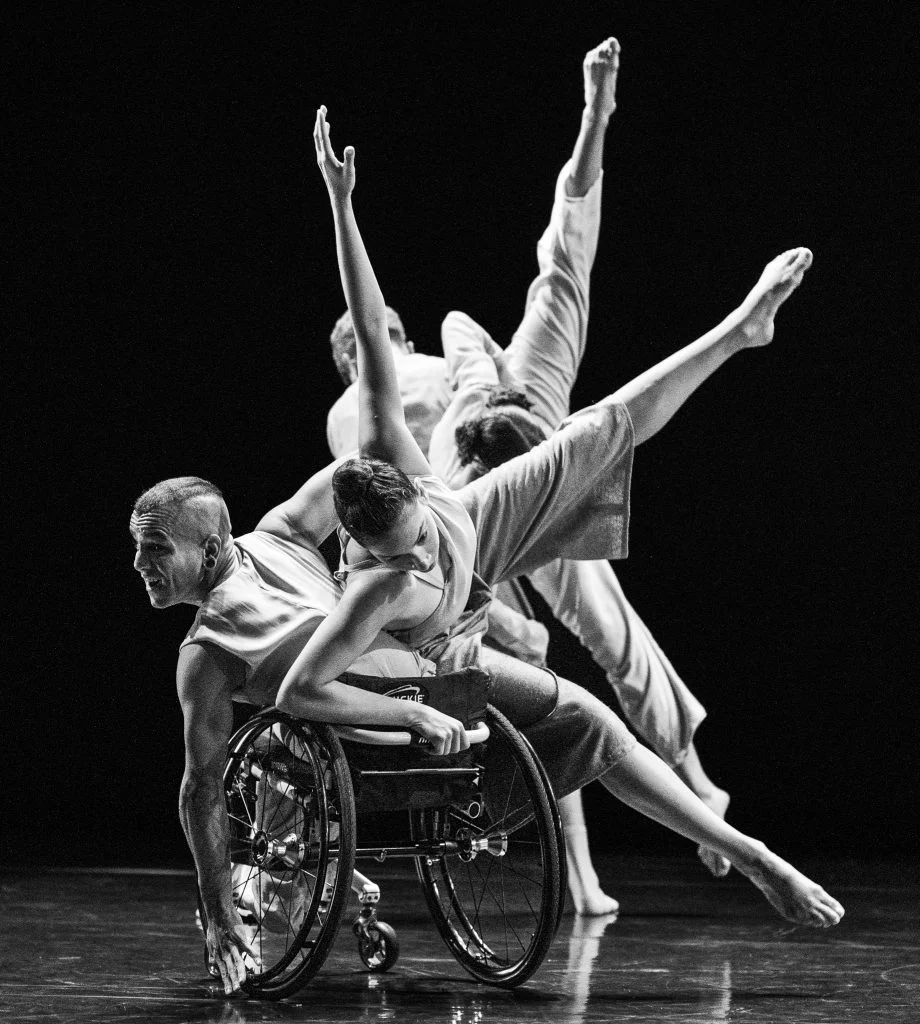 In a black and white photo, Janpi tilts their torso to the left in their wheelchair, while Zara balances in a side tilt on Janpi's body & chair with two legs and one arm extended outwards. David and Alaja perform the same movements behind them both.