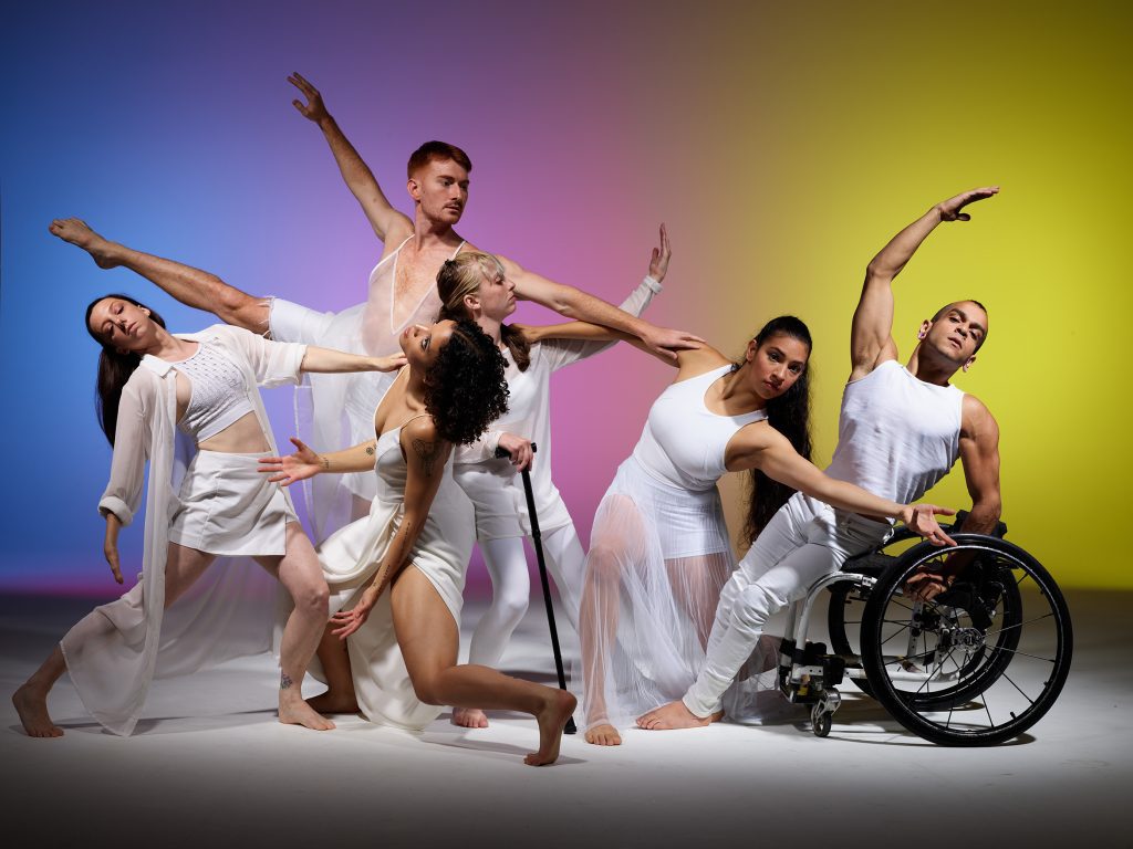 Six disabled and non-disabled dancers pose in white clothes against a rainbow gradient backdrop. The dancers reach, balance and extend various limbs in different directions as they connect to each other. One dancer has a limb difference, another is a wheelchair user and another is a cane user.