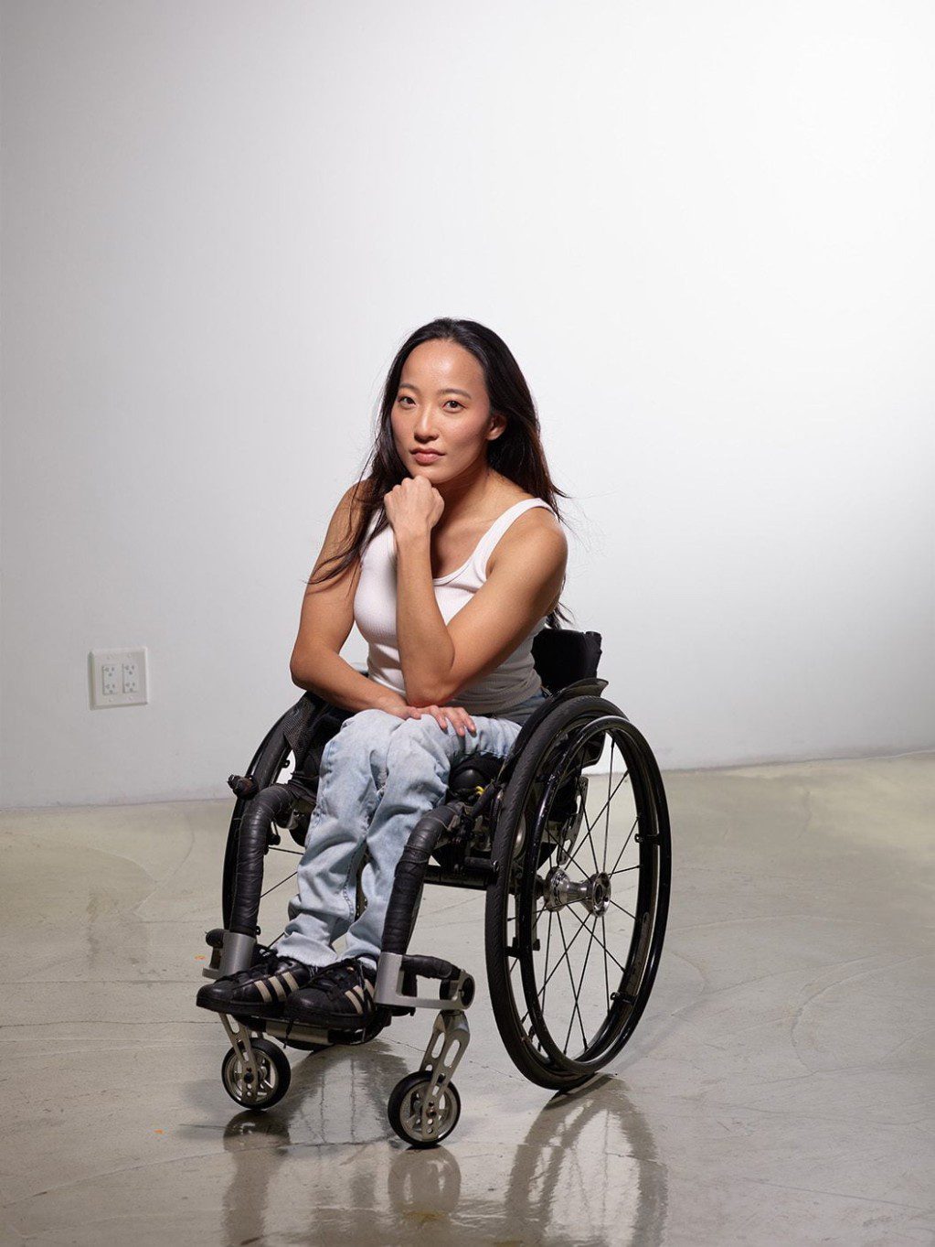 Julie poses for the camera. She is a woman of Japanese descent from Brazil, and is a wheelchair user. She has long black hair and wears a white tank top and blue jeans.