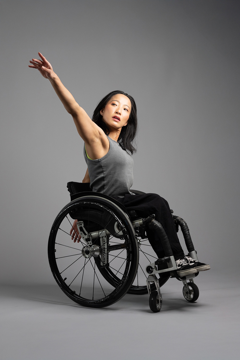 2025AXIS_RobSuguitan_Julie_Portrait Julie lifts one upper limb with elegance in a full body portrait. She is of Asian/Brazilian descent and has dark short hair and brown eyes. She is a wheelchair user and is wearing a grey shirt and black pants.