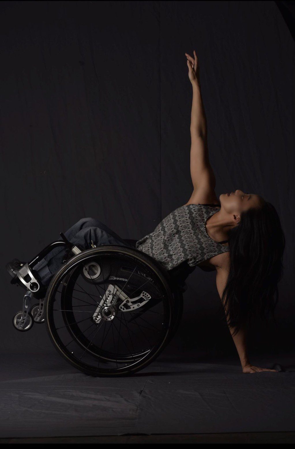 Julie leans back in her manual wheelchair in a wheelie, supporting herself with one hand as she reaches her other arm up to the sky. She poses against a black background, wearing a grey top.