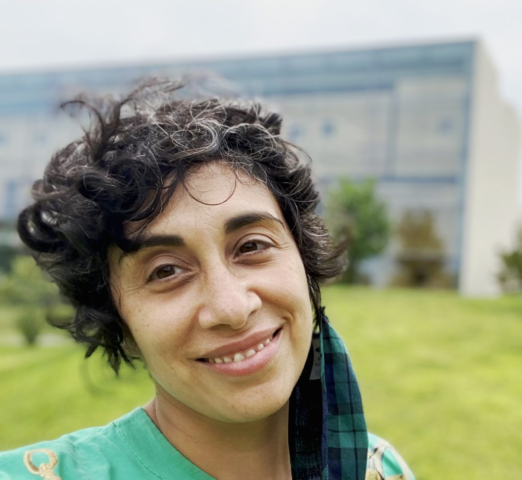A brightly-lit photo of Larissa from the shoulders up. Larissa has light brown skin and short, curly black hair that curls around their foreheard and ears. They are smiling at the camera with a soft expression. They are wearing a teal shirt with a yellow design,and a green mask with a plaid design hangs off of their left ear. In the background, unfocused, is a sprawling green lawn with scattered trees and a rectangular building.