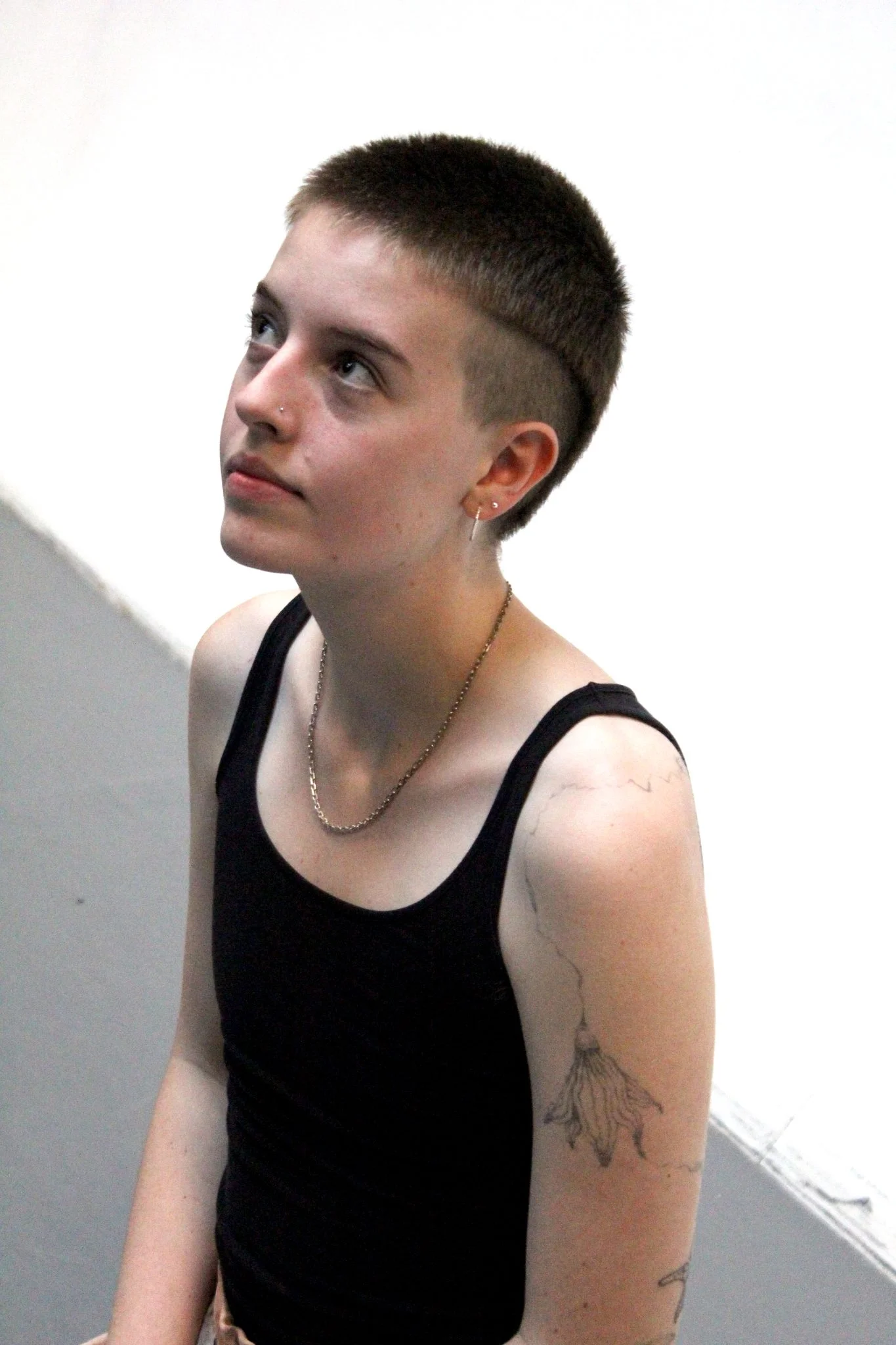 Sammie, a white person in their early 20s, is kneeling on a grey floor with a white wall behind them. They are looking slightly up and to the left.