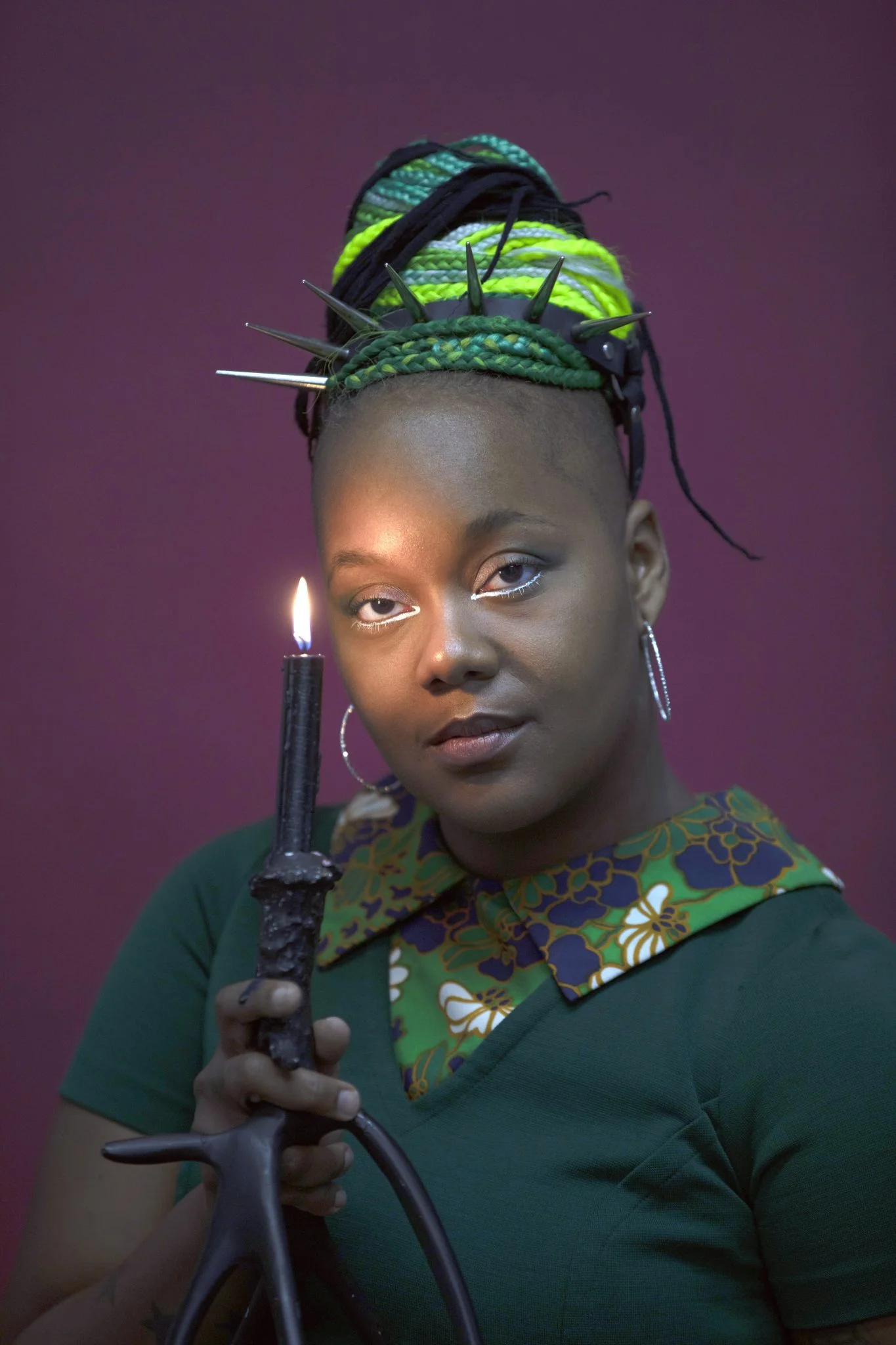 Saira - a queer person with warm brown skin and a green bun - leans one eye toward a candle.