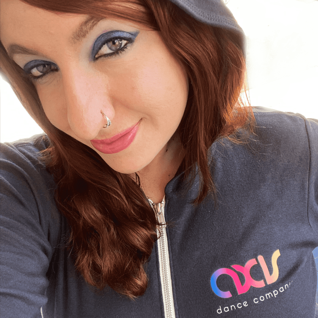 Tashi is white-presenting person with green eyes and wavy dark red hair that reaches slightly past her shoulders. She smiles at the camera and wears a purple eyeshadow and charcoal grey hoodie with the AXIS logo printed on the front left side.